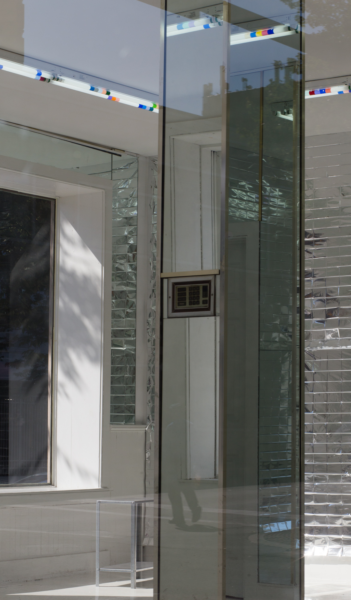 A mirrored column stands in a room, reflecting fluorescent lights wrapped in colored plastic near the top, and a small panel of buttons at its middle