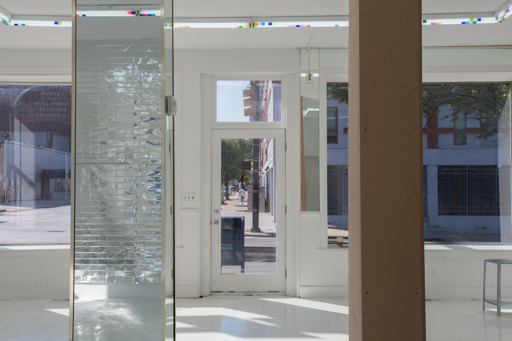 Looking from the back of the room out the storefront windows to the street, the view is intersected by two columns--one is masonite, the other is mirrored and reflects the mylar hanging on the wall behind. Across the street in the distance, a man with a cane in a white shirt walks down the sidewalk