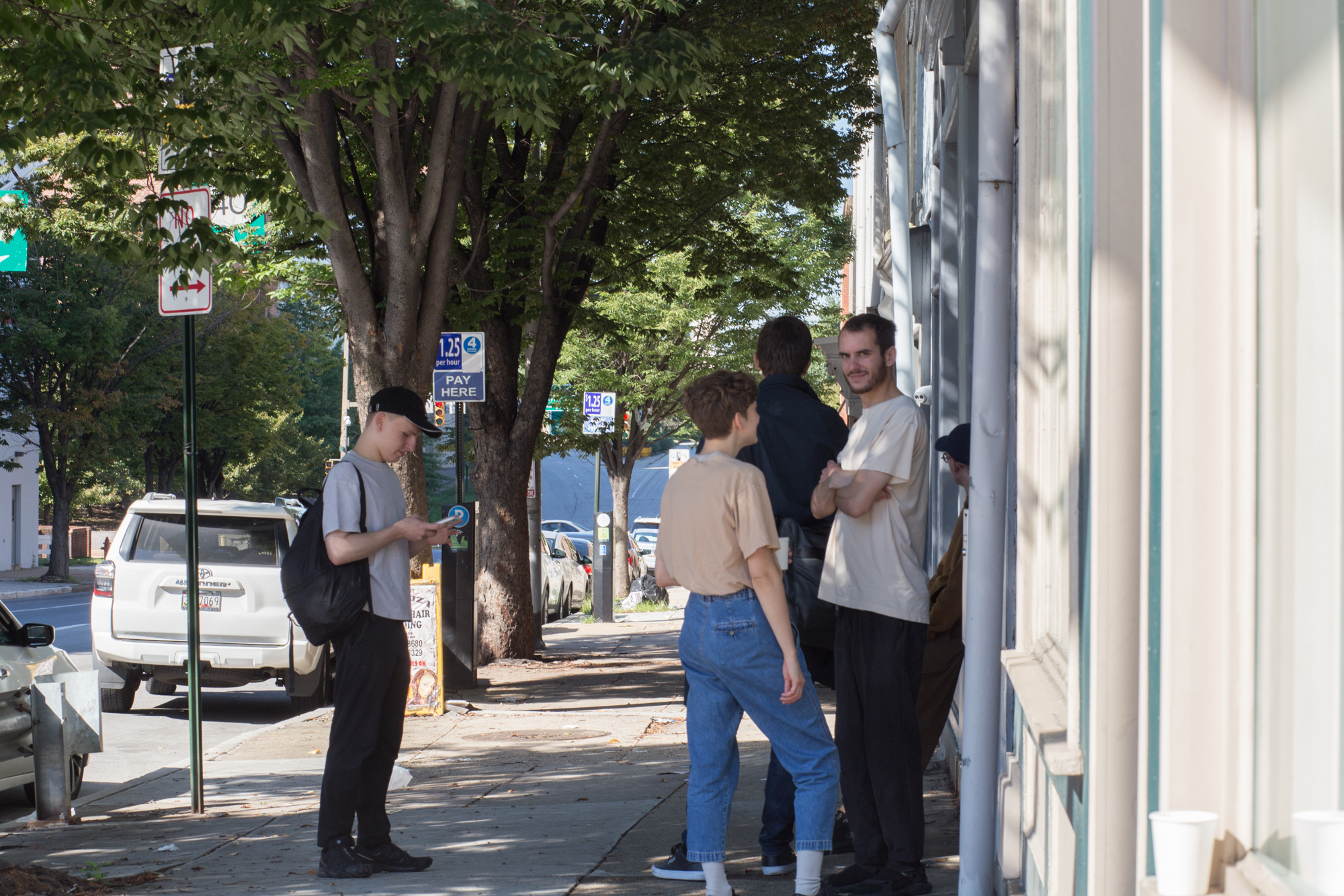 Five people stand on a sidewalk lined with street signs and parking meters. One stands apart from the others, looking at his phone. Two men in the back look away down the street. Two people in the front are in conversation, although one of them looks up at the camera with his arms crossed, smiling