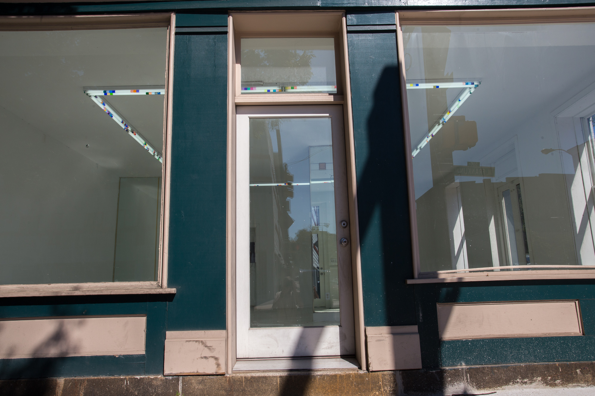 Slightly skewed view of a storefront with glass door and large windows. Inside is a square formation of fluorescent lights, their bulbs wrapping in small tubes of plastic in different colors