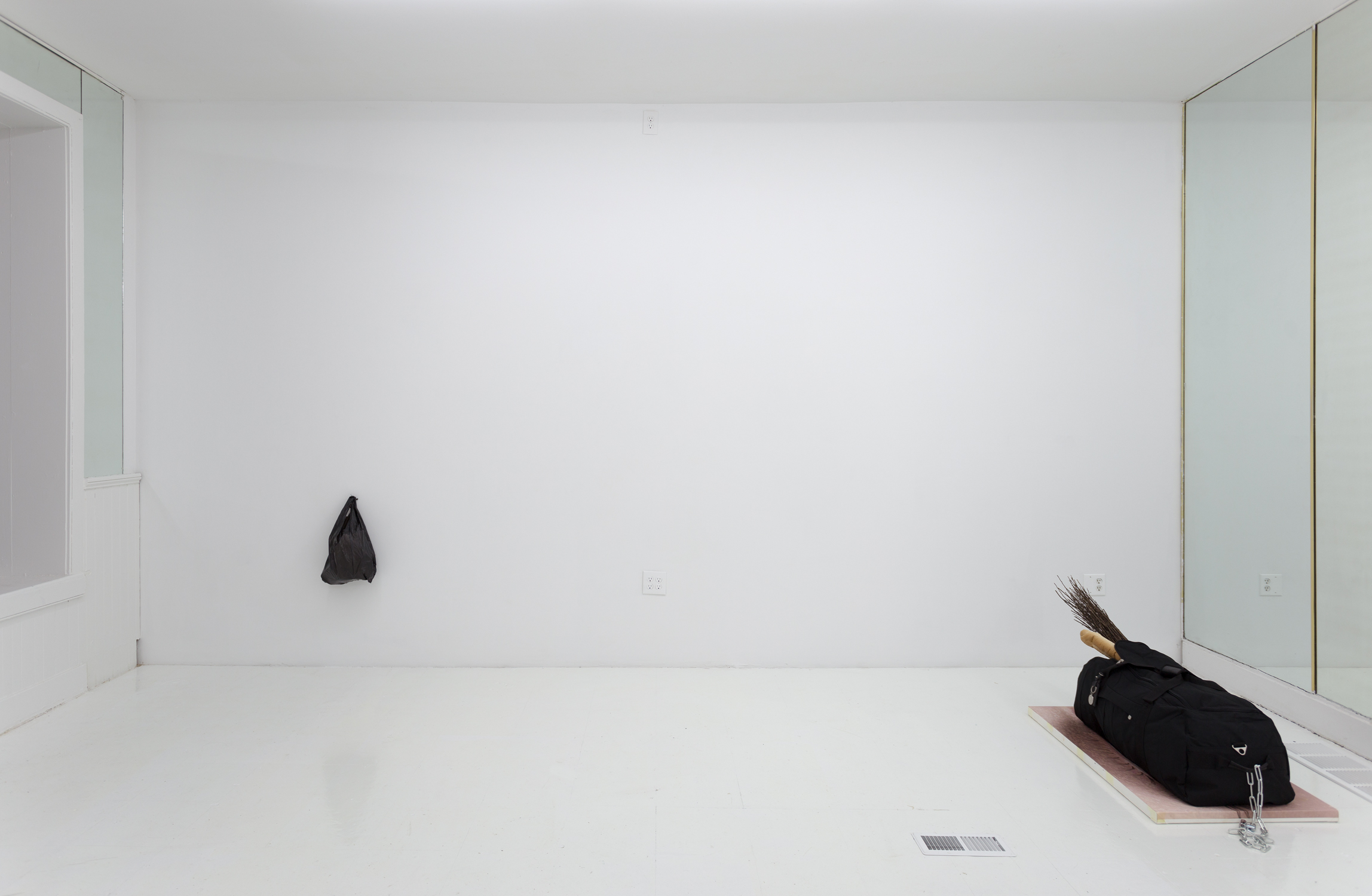 A black plastic bag hangs on a wall to the lower left; on the right, a black duffel bag sits on top of a painting on the floor