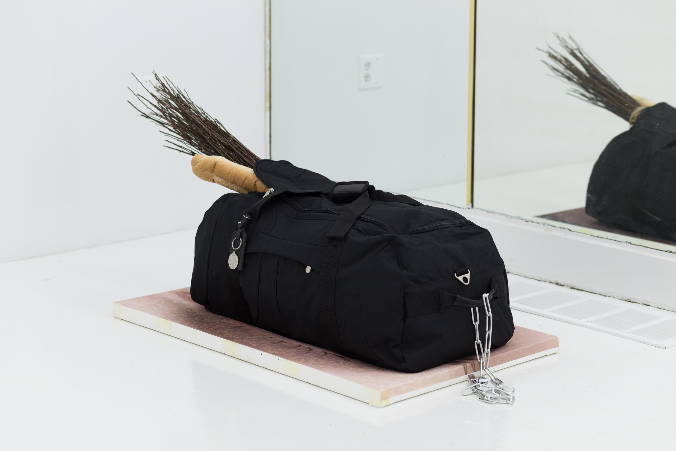A black duffel bag sits atop a painting of an eye. There is a chain attached to the handle; a baguette and cluster of branches sticks out of the bag