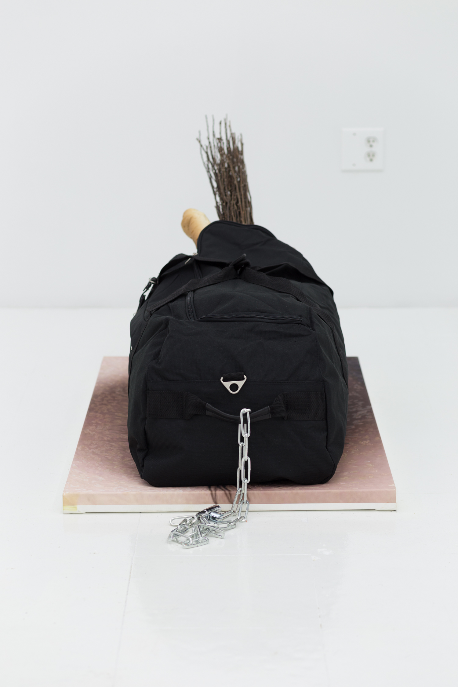 A black duffel bag sits atop a painting of an eye. There is a chain attached to the handle; a baguette and cluster of branches sticks out of the bag