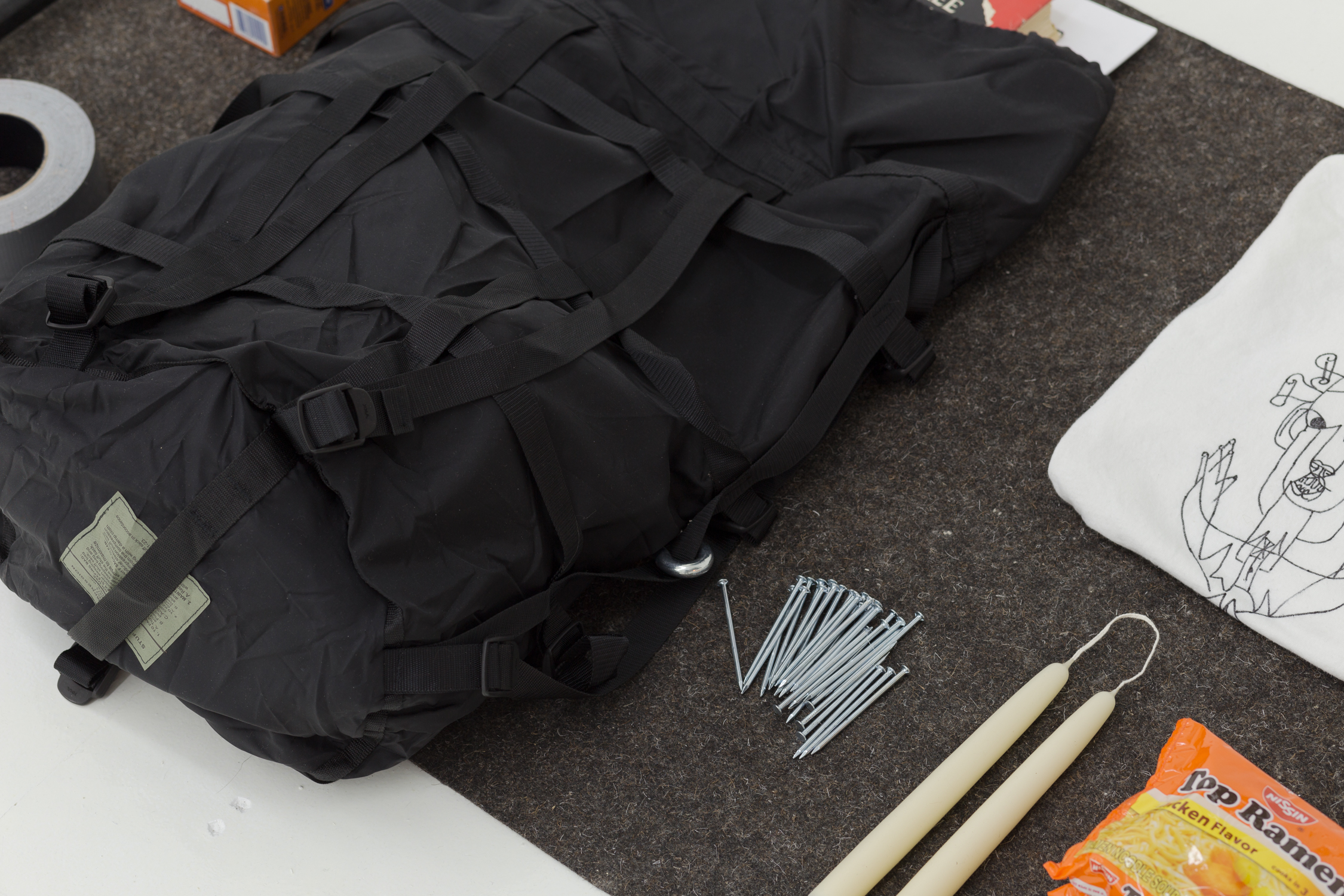 Items on gray felt: black backpack, a pile of nails, a folded t-shirt, two candles, and a package of Top Ramen