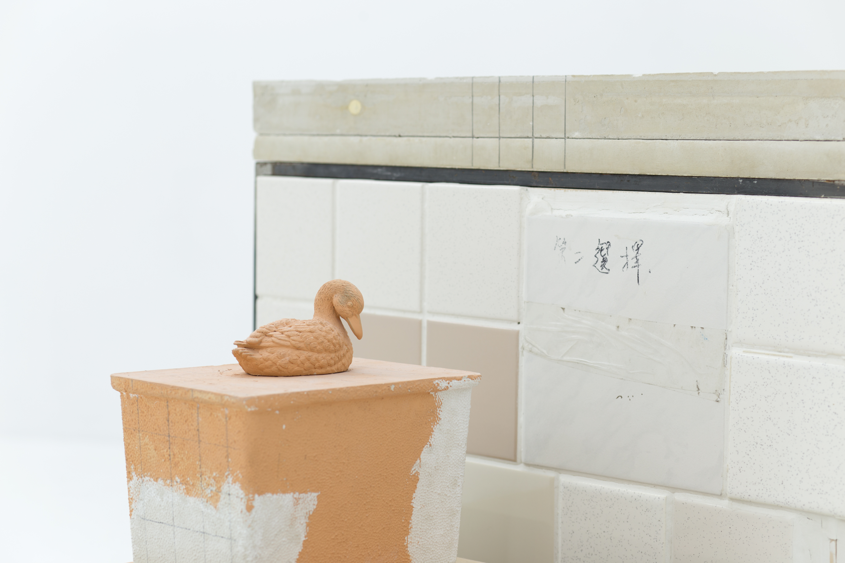 A sculpture consisting of several tile boxes on a wooden pedestal; one is topped with a ceramic duck