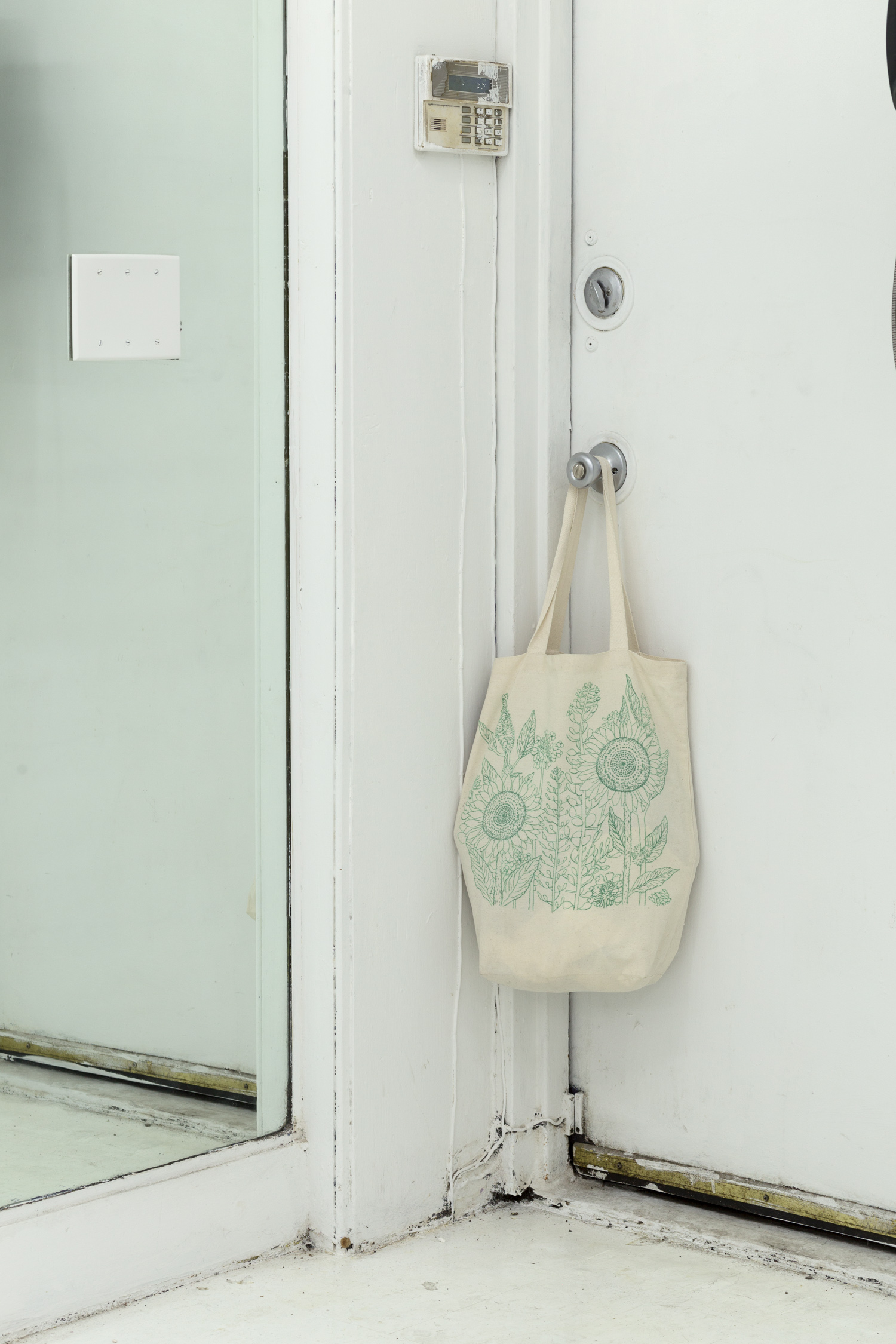 A canvas tote with a drawing of sunflowers in green pen hangs from a doorknob