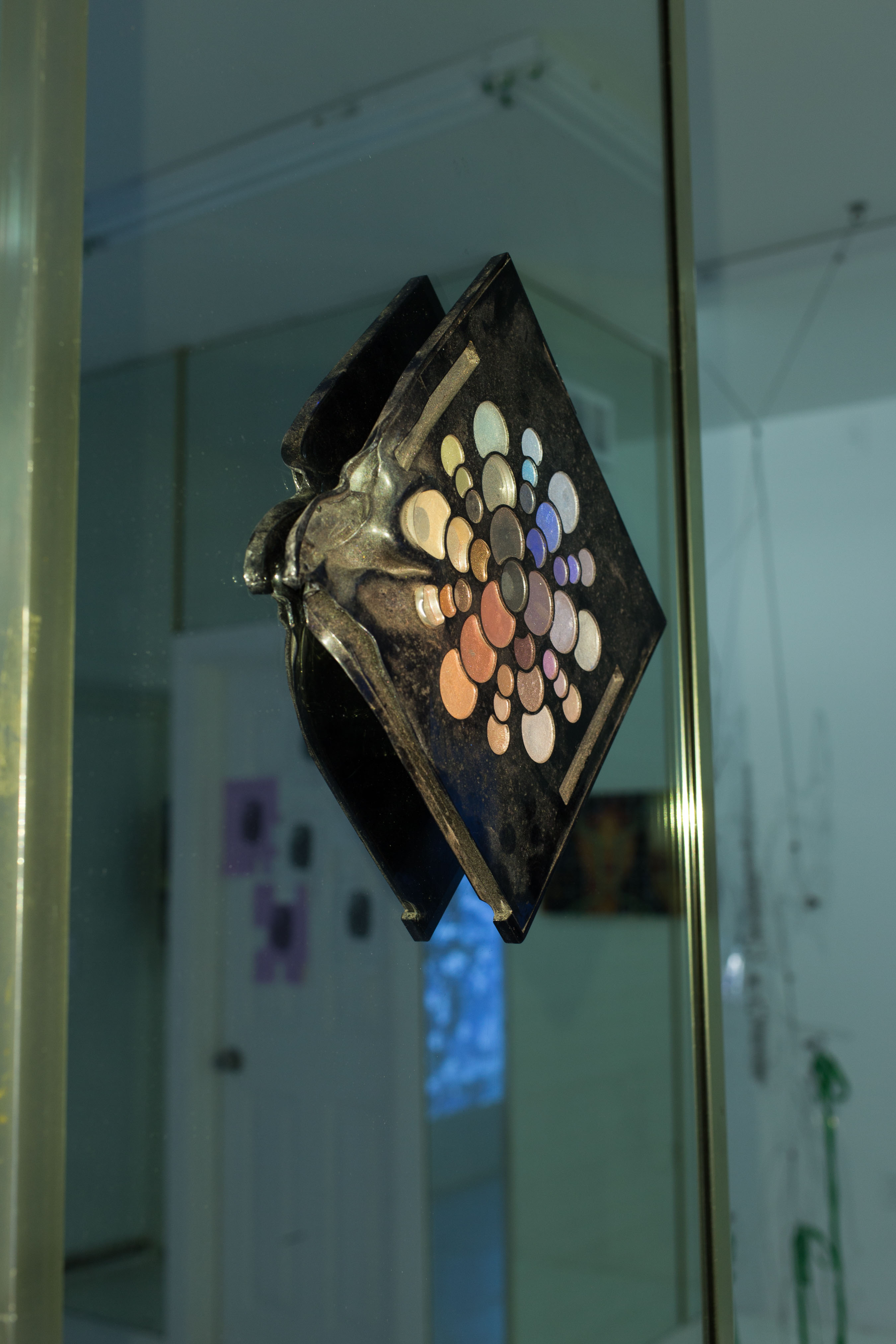 A piece by Dese Escobar: a makeup palette hangs on a mirror; one corner is melted and twisted