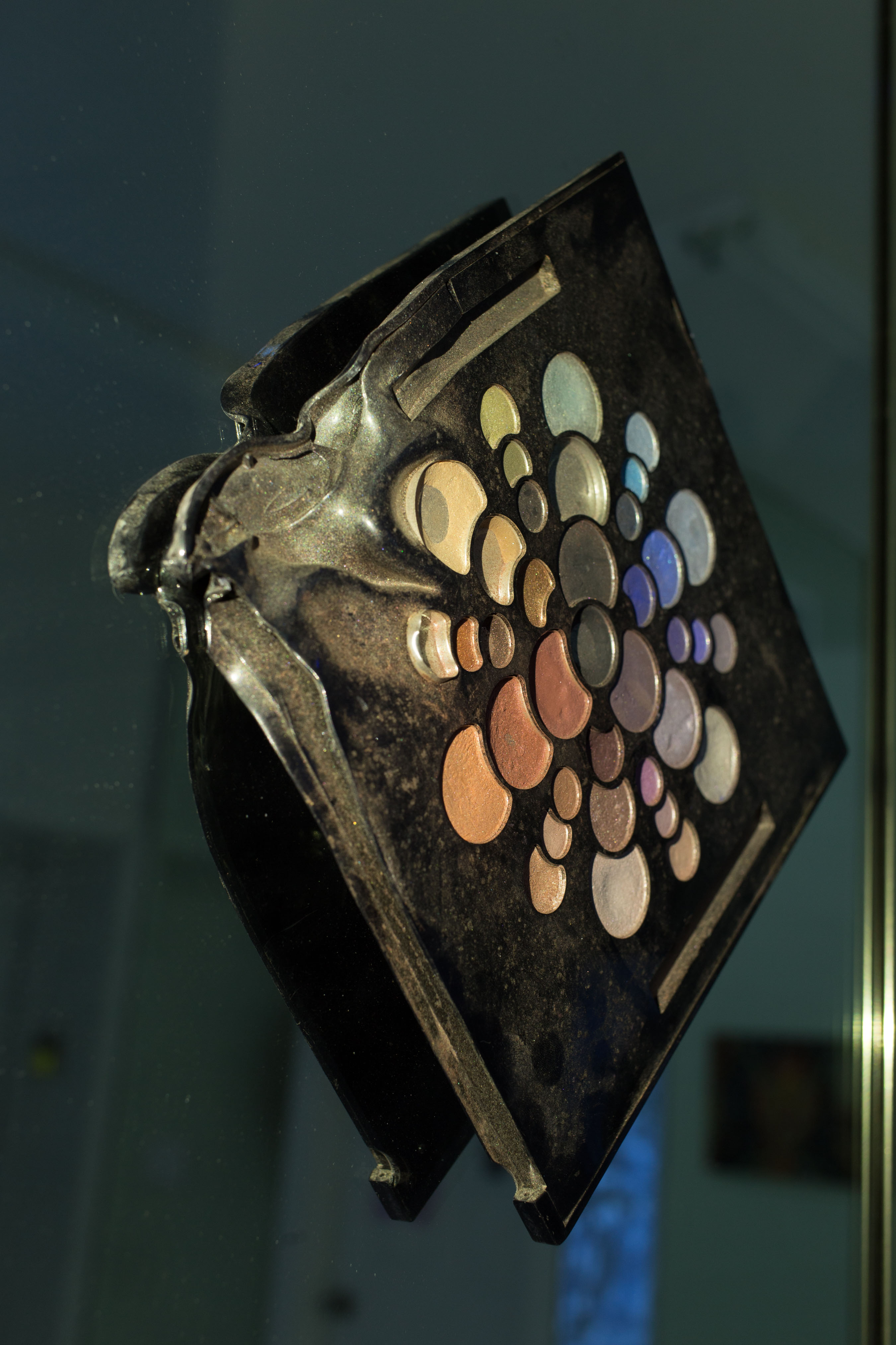 A piece by Dese Escobar: a makeup palette hangs on a mirror; one corner is melted and twisted