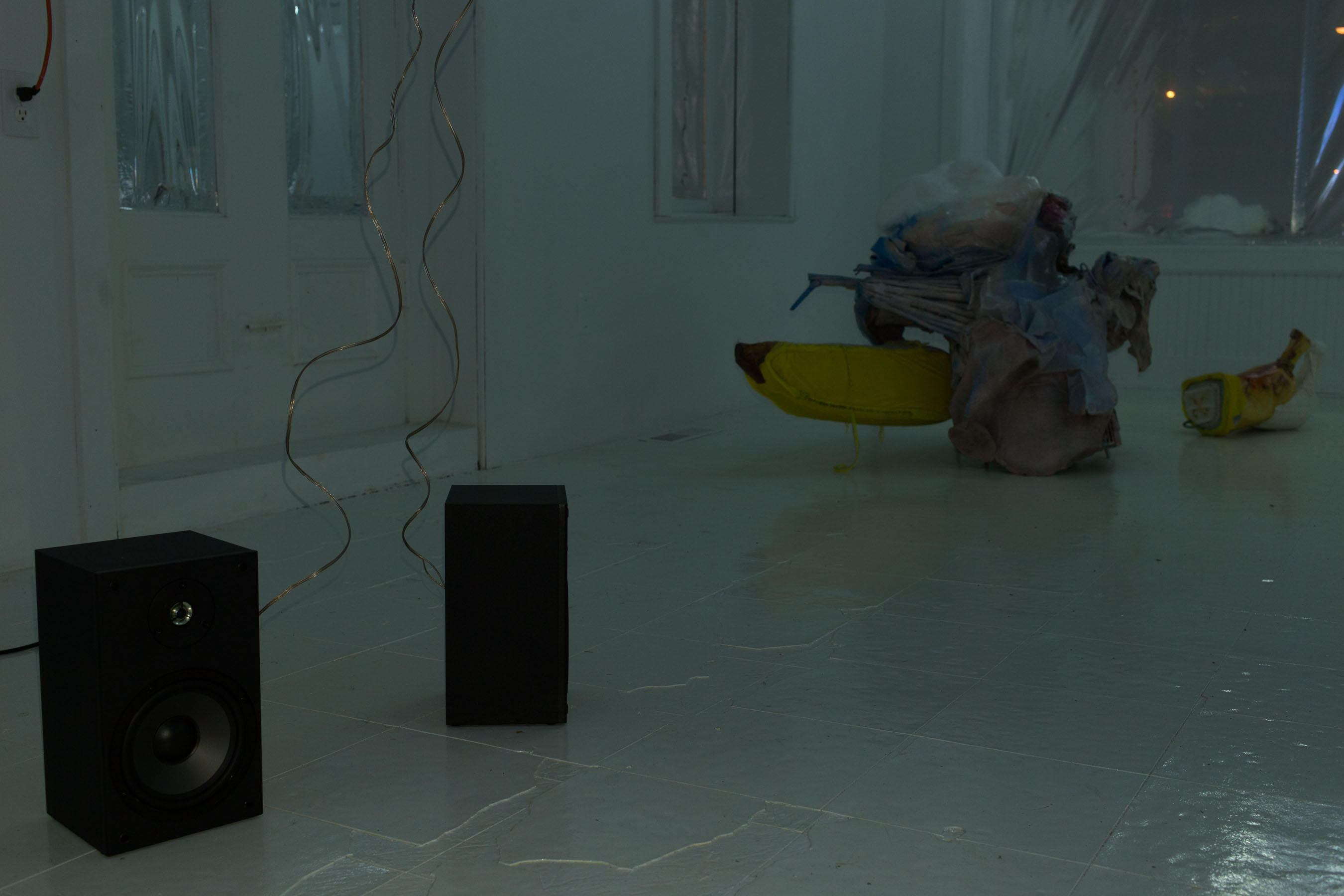 Two speakers sit in space, their copper speaker wire trailing up to the ceiling. In the background sits a sculpture by Danica Barboza