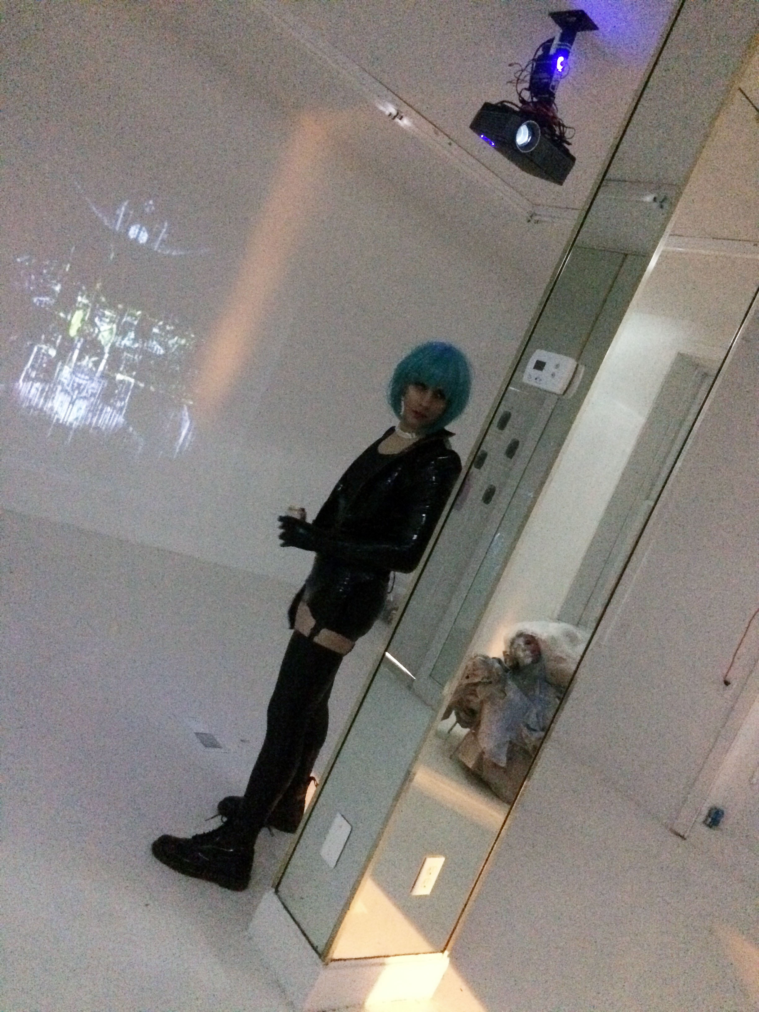 A person in black latex and a blue wig stands beside a mirrored column